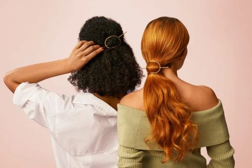 Two women with different hair colors stand close together showcasing gold hairpins in a studio with a soft pink background.