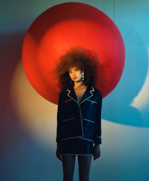 Young woman in stylish suit poses against a backdrop with a large red circle, creating a fashion-forward, artistic portrait.