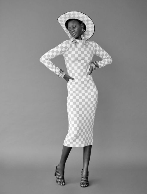 A black and white studio shot of a tall woman in a checkerboard dress, matching hat, and high heels against a neutral background.