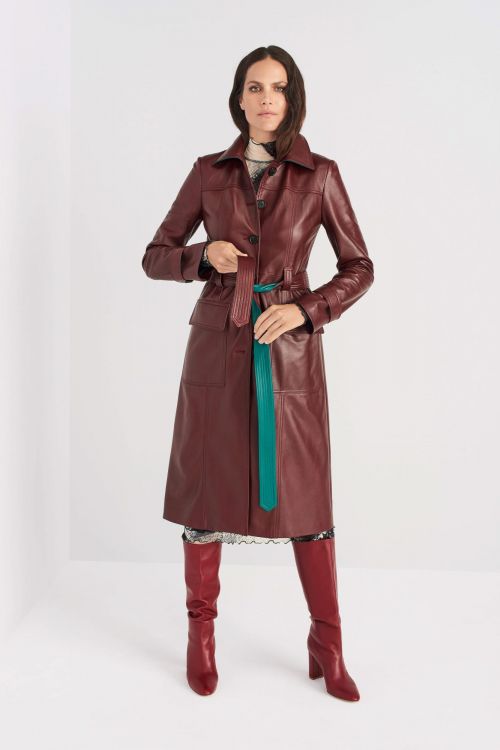 A stylish adult woman poses in a studio wearing a red leather coat, a patterned dress, and matching heeled boots against a clean white backdrop.