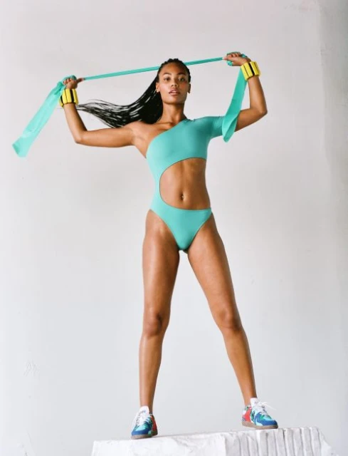 Athletic woman in teal one-piece swimsuit standing on a white surface with a teal resistance band and yellow wrist weights.