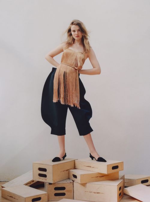 A woman with blonde hair poses on a stack of wood boxes wearing a sleeveless tassel top and cropped balloon pants in a studio setting.