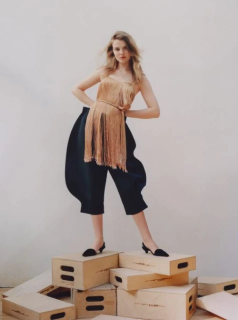 A woman with blonde hair poses on a stack of wood boxes wearing a sleeveless tassel top and cropped balloon pants in a studio setting.