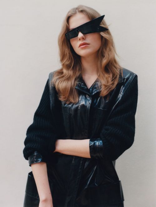 A woman in a black jacket and unique sunglasses poses against a neutral background, creating a high-fashion editorial portrait.
