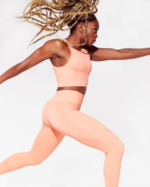 Athletic woman in a peach-colored sports bra and leggings leaps against a bright white backdrop in a fitness-focused shot.