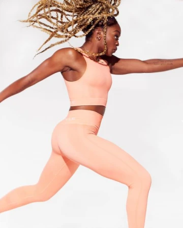 Athletic woman in a peach-colored sports bra and leggings leaps against a bright white backdrop in a fitness-focused shot.