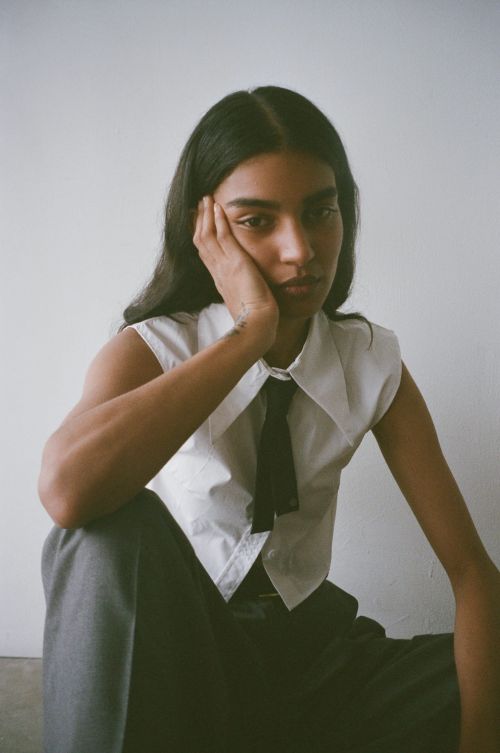 Portrait of a young adult woman in a sleeveless white shirt and grey pants, with dark hair, posing in front of a plain background.