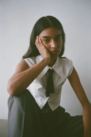 Portrait of a young adult woman in a sleeveless white shirt and grey pants, with dark hair, posing in front of a plain background.