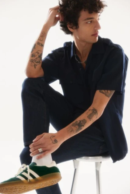 A curly-haired adult male sitting on a stool in a navy shirt and pants, green and white tennis shoes with tattoos on forearms in studio.