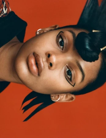 Close-up portrait of a woman with dramatic black eyeliner, styled dark hair, and hoop earrings against an orange background.