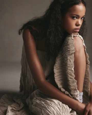 A young woman with curly dark hair sitting on the floor wearing a textured neutral-colored dress with ruffled edges, styled by Calvy Click.