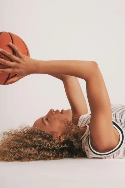Adult female lying on her back, holding a basketball with both hands in a light-colored studio environment. She is wearing a mesh jersey.