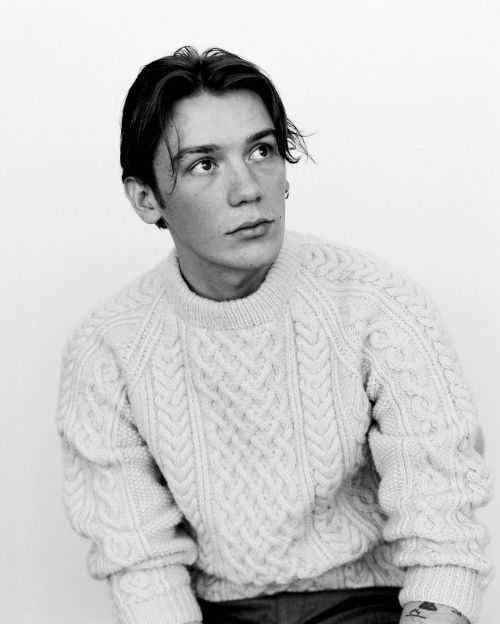 Monochrome portrait of a young man with center-parted hair wearing a cream cable-knit sweater against a white seamless background
