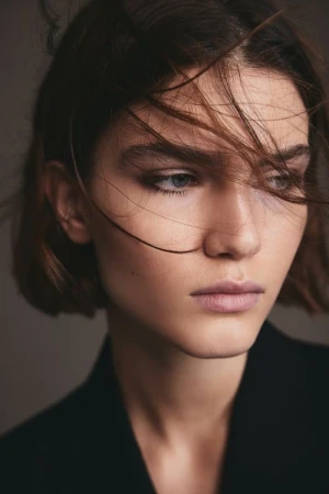 Close-up portrait of a woman with short dark hair blowing across her face and wearing a black jacket against a dark background.
