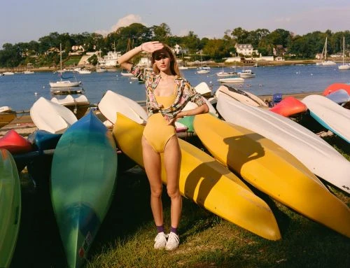 A woman in a yellow swimsuit poses with kayaks on a grassy area by a bay with boats, houses, and trees in the background on a sunny day.
