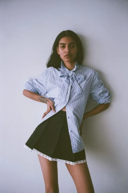 Young adult female poses indoors wearing a light blue striped blouse with a bow tie and a black mini skirt with white lace trim.