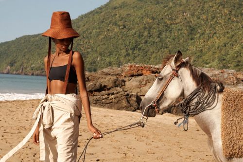 Woman in sun hat poses with horse on sandy beach with coastal vegetation and blue ocean waters in background on a sunny day.