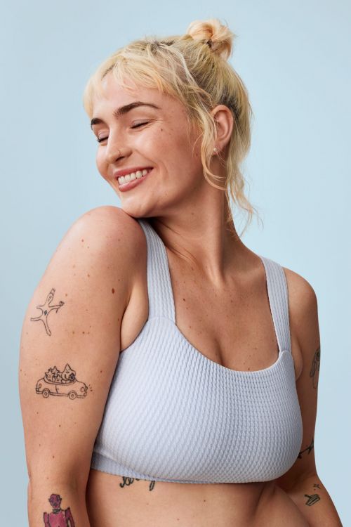Smiling woman with blonde hair in a casual top against a light blue background, showcasing arm tattoos. Beauty and fashion portrait.