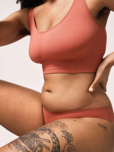 Body positive portrait of a young woman in a ribbed top and underwear, posing in front of a neutral background, showing her natural figure.