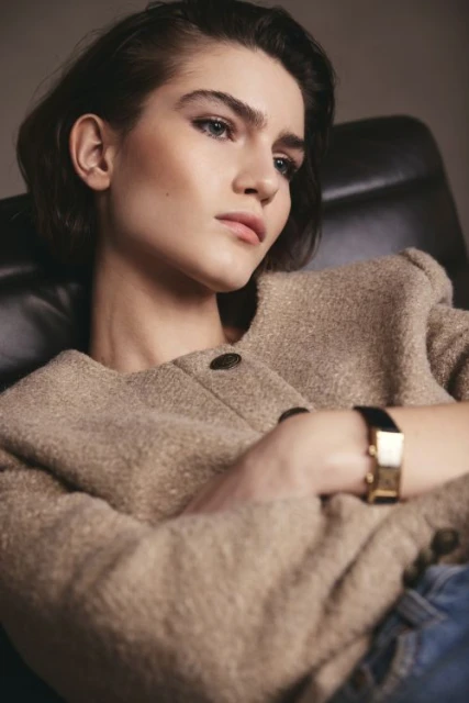 Portrait of a brunette adult female in a beige wool jacket and jeans, seated indoors, with soft lighting and a shallow depth of field.