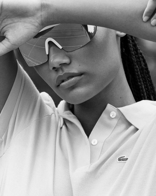 Black and white portrait of a woman wearing sporty sunglasses and a buttoned polo shirt, her hands are raised to her head.