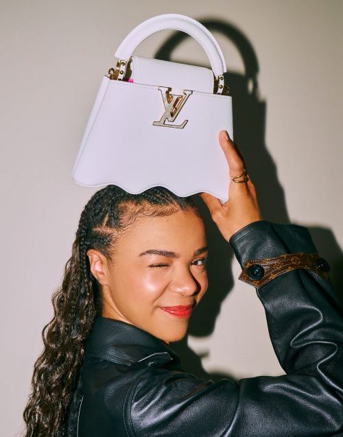 A fashionable adult female winks at the camera while posing with a white designer purse that features a gold logo and handle.
