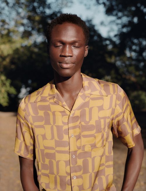 Portrait of a Black man with eyes closed, wearing a patterned short-sleeved shirt, standing outdoors surrounded by trees on a sunny day.