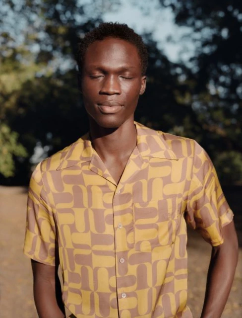 Portrait of a Black man with eyes closed, wearing a patterned short-sleeved shirt, standing outdoors surrounded by trees on a sunny day.