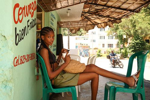 Young woman with braided hair relaxing outdoors in a chair at a roadside cafe, holding a drink, with a motorcycle passing by.