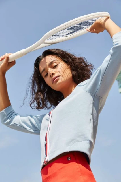 Woman in blue sweater and red skirt poses with tennis racket against blue sky. Sporty fashion shoot with natural light.