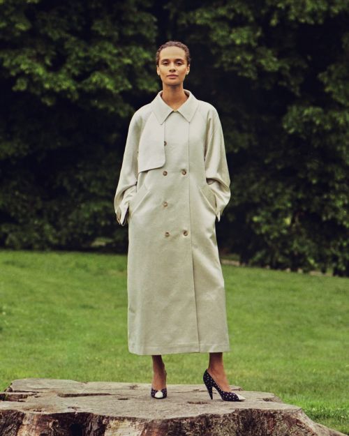 A woman in a long beige coat poses outdoors on a tree stump, with a green lawn and trees behind her, in a full shot.