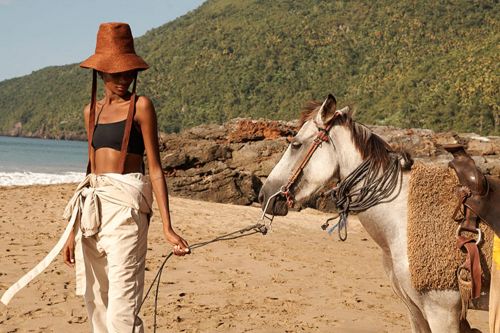 Model wearing a wide-brimmed brown hat and beige trousers on a sandy beach holding reins of a horse