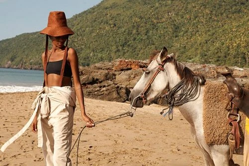 Model wearing a wide-brimmed brown hat and beige trousers on a sandy beach holding reins of a horse