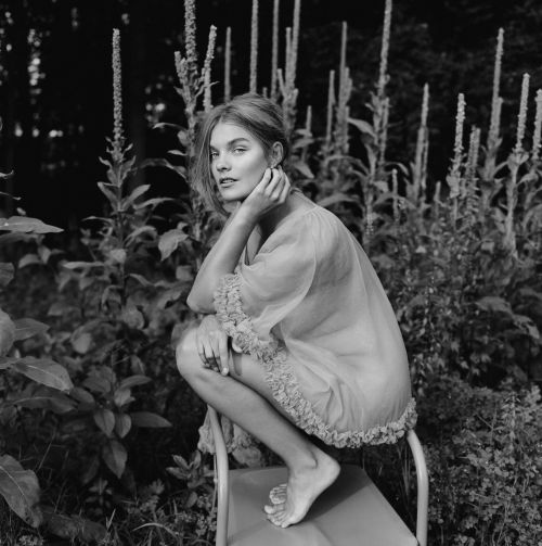 Monochrome portrait of a barefoot, fair-skinned young woman with long hair, crouching on a metal frame amidst lush foliage in a garden setting.