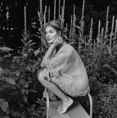 Monochrome portrait of a barefoot, fair-skinned young woman with long hair, crouching on a metal frame amidst lush foliage in a garden setting.