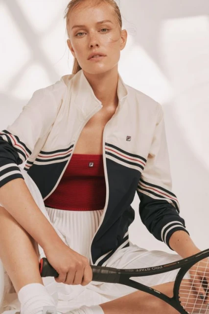 Young woman in white tennis outfit with Fila jacket holding racket, sitting on a white background with soft lighting, fashion shot.