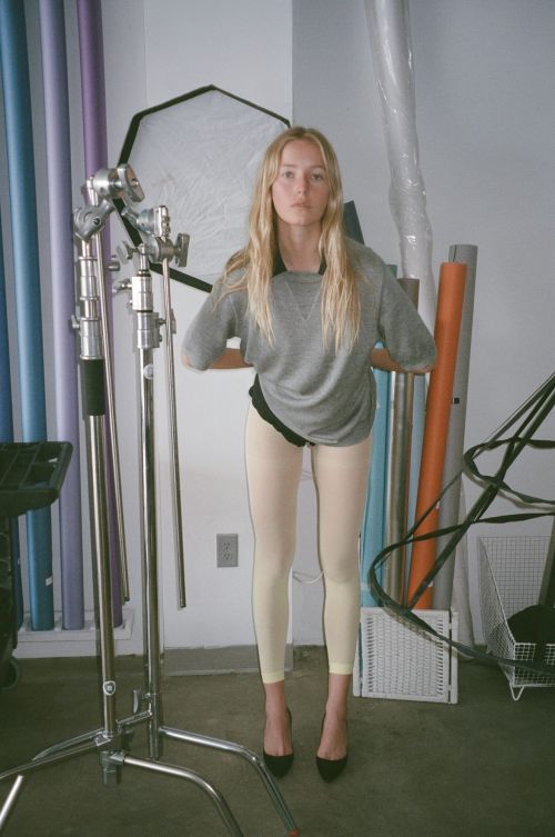 An adult female poses indoors in a photo studio with fair skin, long blonde hair, a grey sweatshirt, light leggings, and black heels.