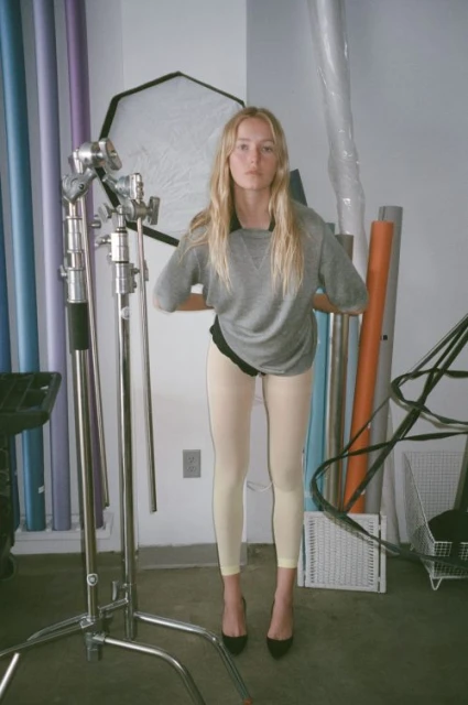 An adult female poses indoors in a photo studio with fair skin, long blonde hair, a grey sweatshirt, light leggings, and black heels.