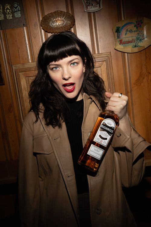 Woman holding bottle of whiskey, wearing a tan coat, red lipstick and blunt bangs, standing in front of wood paneled wall.