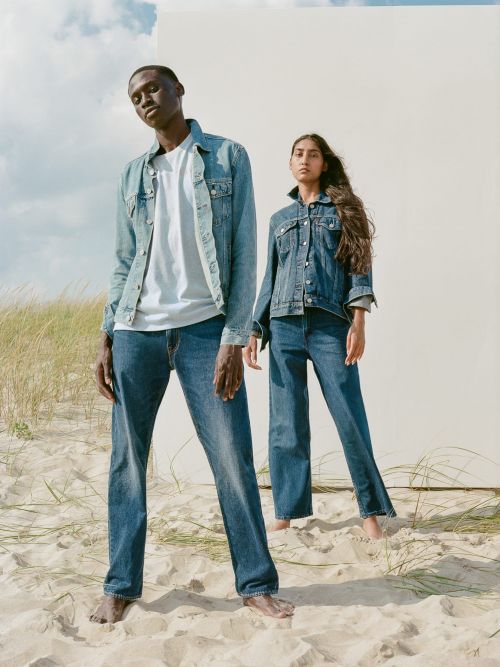 Two models wearing denim outfits standing on a sandy beach. The dark-skinned man on the left is barefoot; the woman has long, wavy hair.