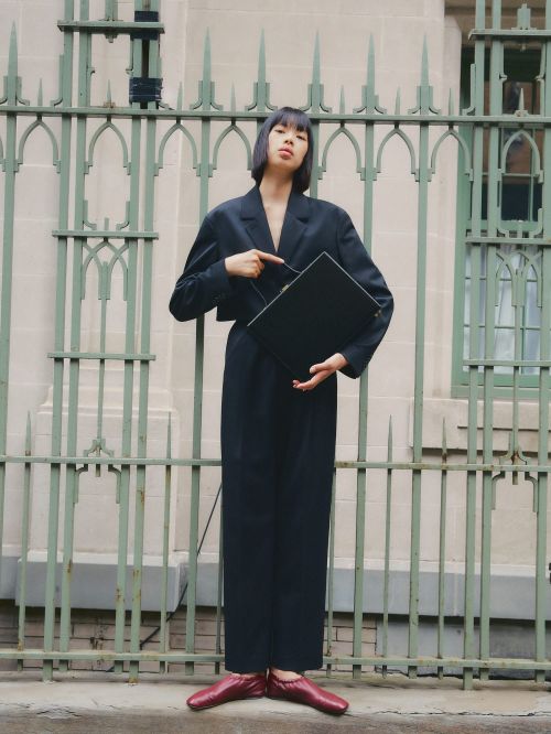 A stylish woman in a navy suit poses confidently outdoors, pointing at a black laptop against an ornate fence backdrop, wearing red shoes.