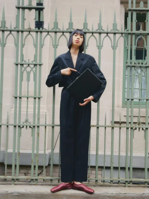 A stylish woman in a navy suit poses confidently outdoors, pointing at a black laptop against an ornate fence backdrop, wearing red shoes.