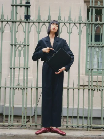 A stylish woman in a navy suit poses confidently outdoors, pointing at a black laptop against an ornate fence backdrop, wearing red shoes.