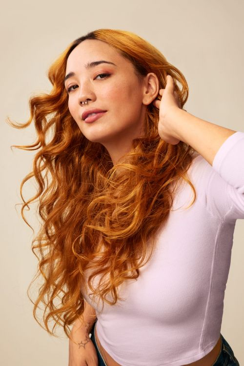 Portrait of a young adult woman with curly auburn hair, wearing a light purple long-sleeve shirt, posing in a studio with a neutral background.