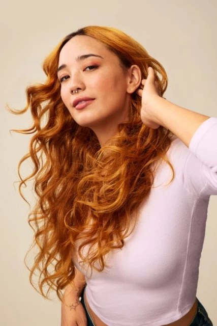 Portrait of a young adult woman with curly auburn hair, wearing a light purple long-sleeve shirt, posing in a studio with a neutral background.