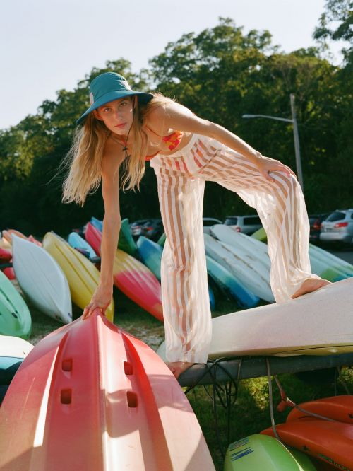 A young woman in a sun hat and striped pants stands on a stack of colorful kayaks in an outdoor setting on a sunny day.