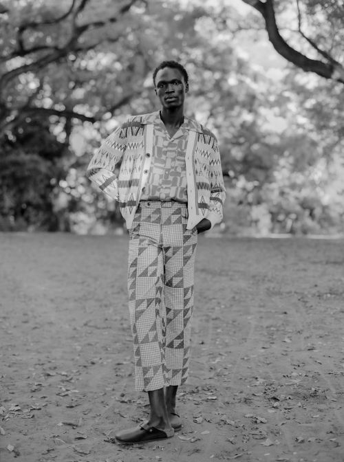 Black and white portrait of an African American man wearing a patterned shirt, cardigan, and cropped pants in an outdoor setting.
