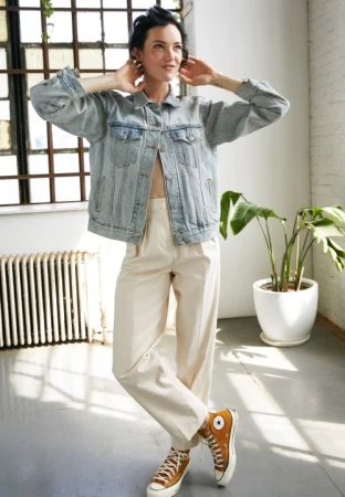 Woman in oversized denim jacket, cream trousers and mustard sneakers posing near a sunny window in New York.