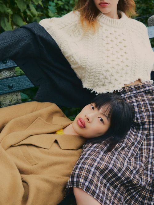 Two women in stylish casual outfits lounge outdoors. One wears a tan coat, while the other has on a cropped sweater and plaid skirt.