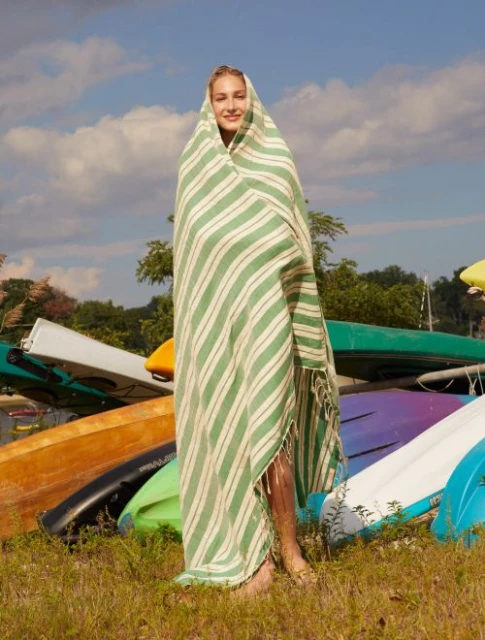 Young fair-skinned woman stands wrapped in a striped green and white beach towel outdoors among upturned kayaks on a sunny day.
